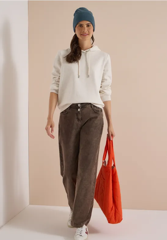 Femme en sweat à capuche blanc, jean marron ample, bonnet bleu et sac rouge devant un fond neutre.