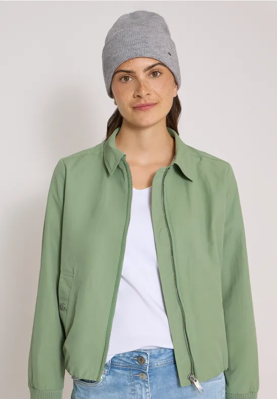 Jeune femme portant un bonnet gris, un t-shirt blanc et une veste verte sur fond uni.