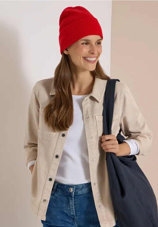 Une femme souriante portant un bonnet rouge, une surchemise beige, un t-shirt blanc et un jean, sac sombre à l’épaule.