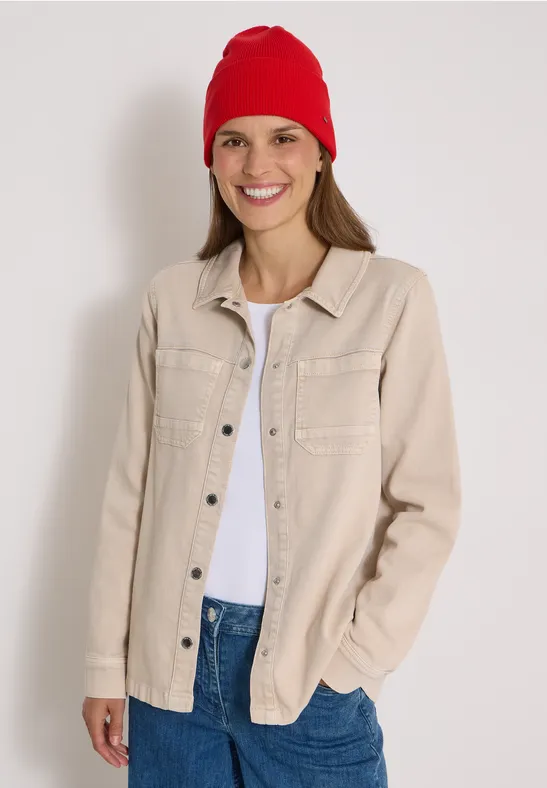 Jeune femme souriante en bonnet rouge, veste beige et t-shirt blanc sur fond clair.