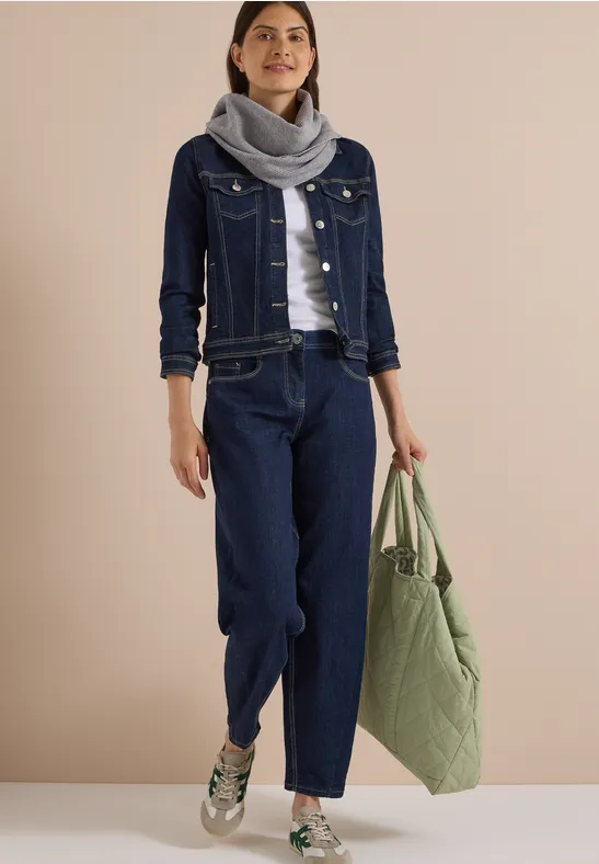 Femme en veste et pantalon en jean bleu foncé, écharpe grise, baskets blanches et grand sac vert.