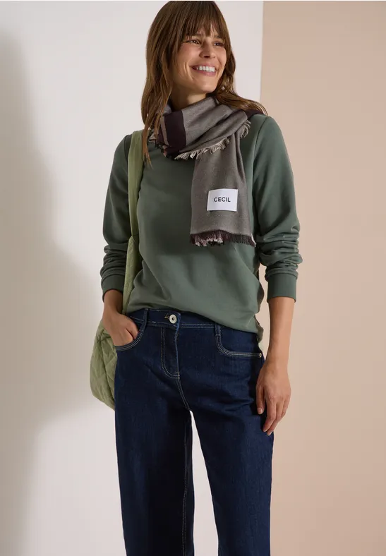 Femme portant un pull vert, une écharpe, un jean et un sac, souriante devant un fond neutre.