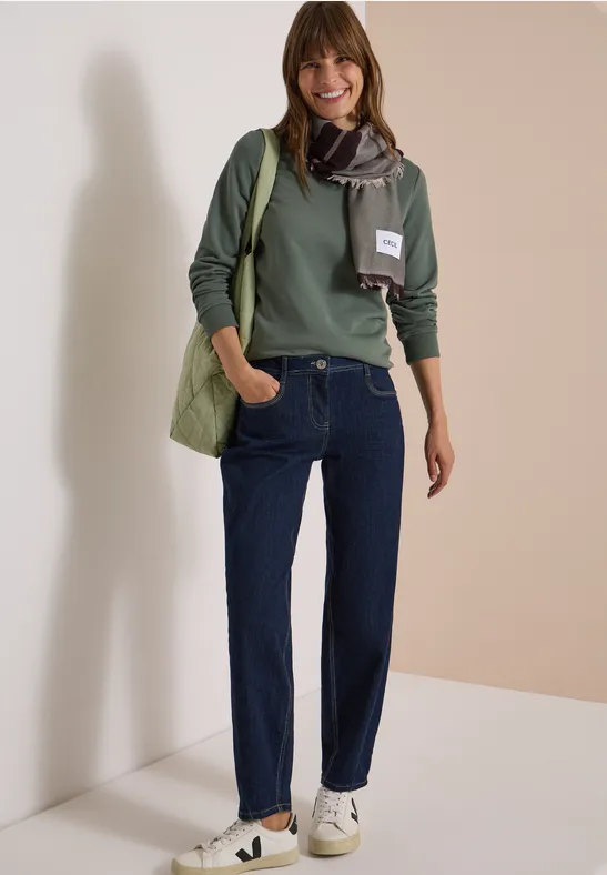 Une femme souriante porte un pull vert, un jean, une écharpe, un sac et des baskets blanches sur un fond neutre.