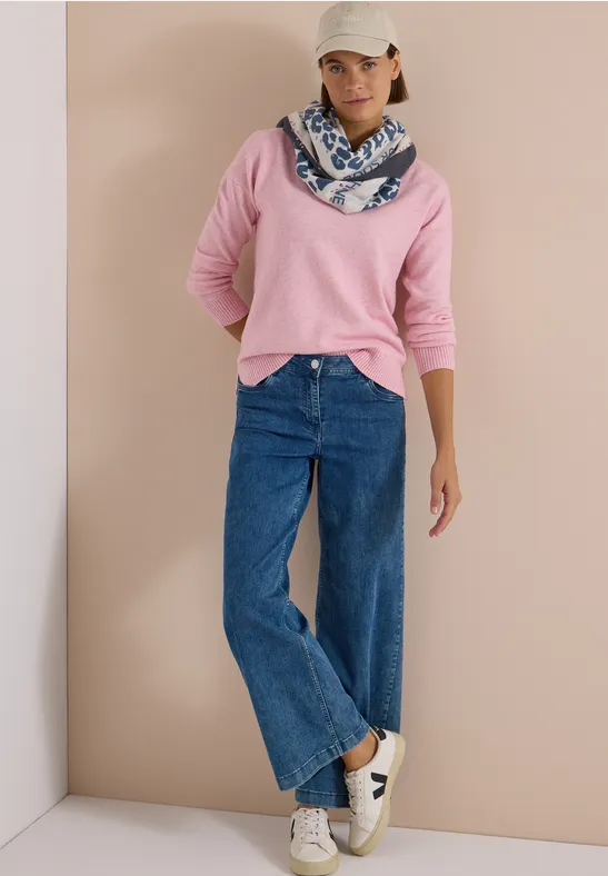 Frau in rosa Pullover, weite blaue Jeans, weißer Cap, gemusterter Schal und weißen Sneakern vor schlichtem Hintergrund.