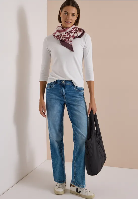 Frau trägt weiße Bluse, blaue Jeans, gemusterten Schal und hält eine schwarze Tasche.