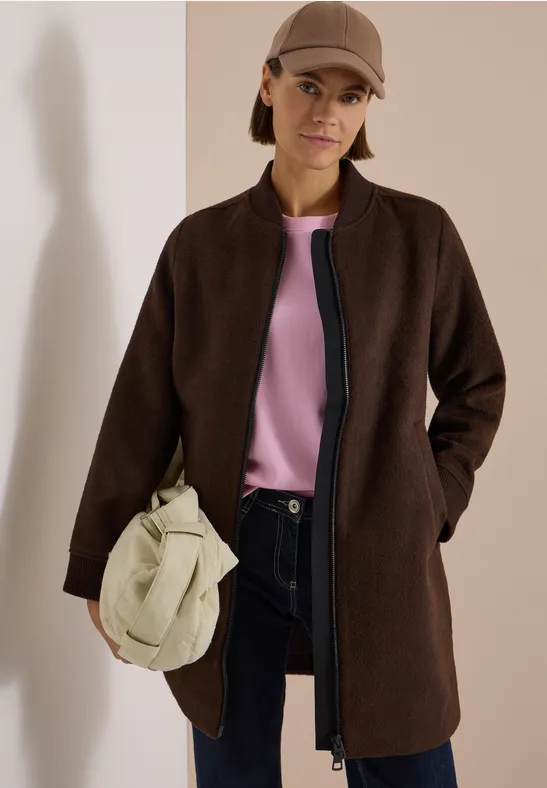Femme en manteau marron, haut rose, pantalon foncé, sac beige et casquette.