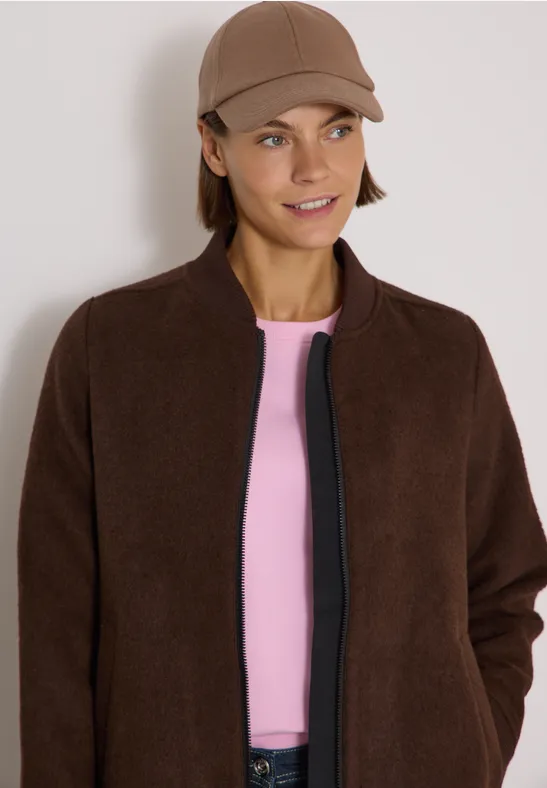 Femme portant une veste marron, t-shirt rose et casquette, souriante.