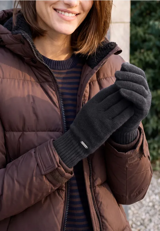 Femme en doudoune marron, pull rayé et gants gris souriant en extérieur.