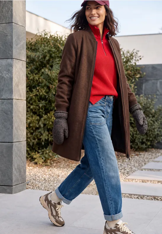 Frau in rotem Pullover, braunem Mantel, Jeans und Handschuhen spaziert im Außenbereich.