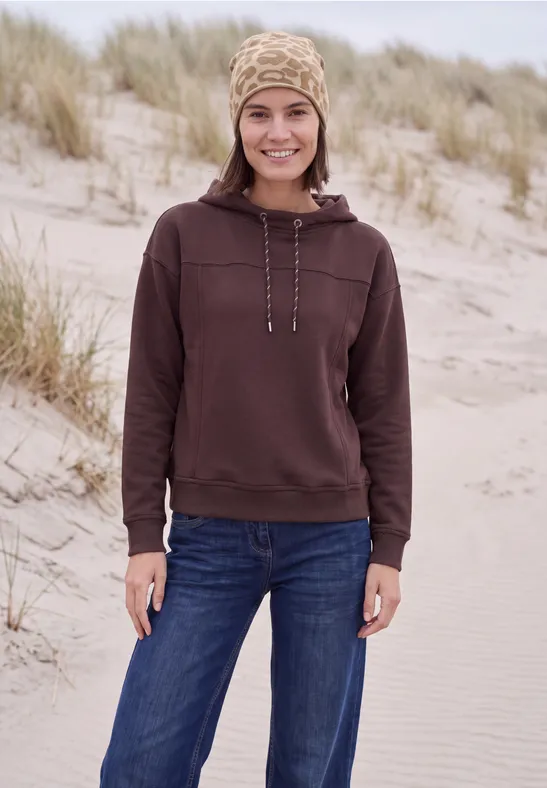 Frau in braunem Hoodie und Jeans steht am Strand vor Dünen und lächelt in die Kamera.