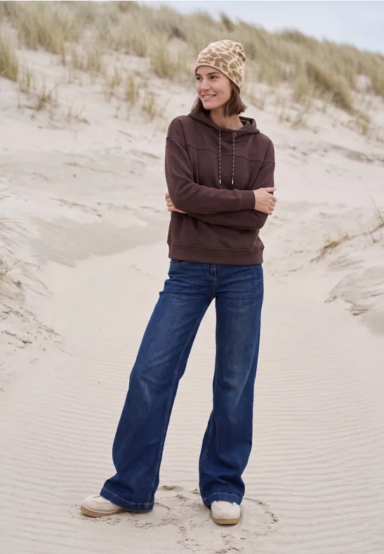 Junge Frau im Hoodie und Strickmütze steht in Dünen am Strand.
