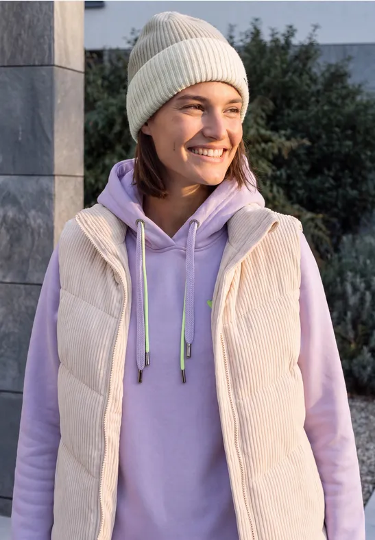 Glimlachende vrouw met beige muts, lichtpaarse hoodie en bodywarmer voor een stenen muur en struiken