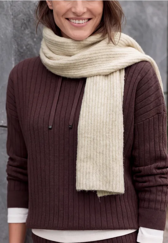 Eine lächelnde Frau in braunem Pullover und cremefarbener Schal vor grauem Hintergrund.