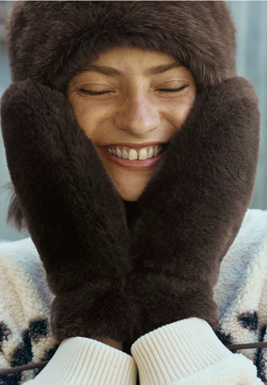 Une femme souriante, emmitouflée dans un bonnet et des moufles en fourrure, se couvre le visage.