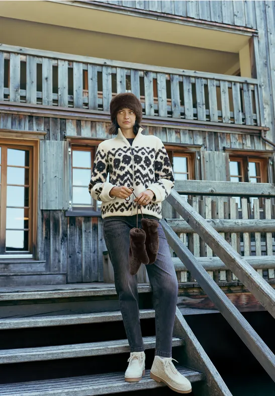 Jeune homme en pull jacquard et bonnet se tient sur un escalier en bois devant un chalet.