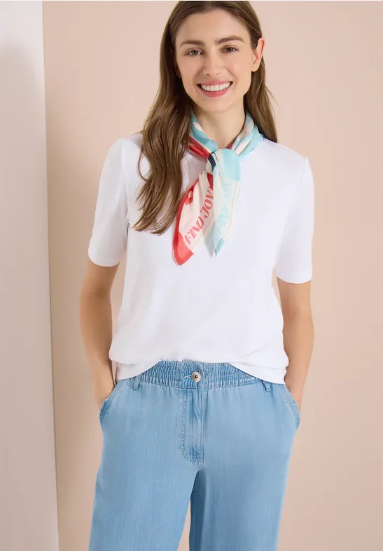 Vrouw met wit T-shirt, lichtblauwe jeans en pastelkleurige sjaal voor een eenvoudige achtergrond.