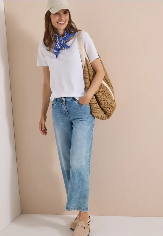 Person mit weißem T-Shirt, blau-weißem Halstuch, blauen Jeans und Strohtasche vor hellem Hintergrund.