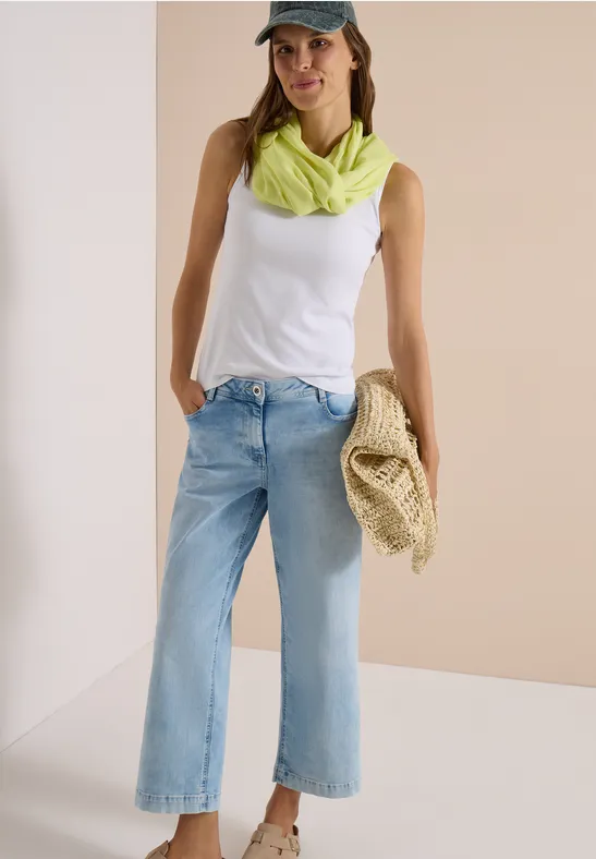 Femme en débardeur blanc, jean clair large, foulard vert néon et casquette, tient un sac en paille.
