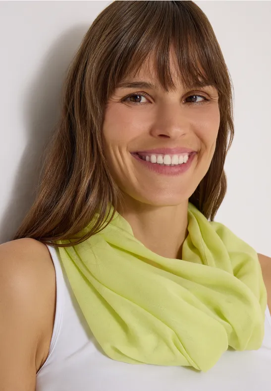 Une femme souriante en débardeur blanc porte un foulard vert clair devant un fond neutre.