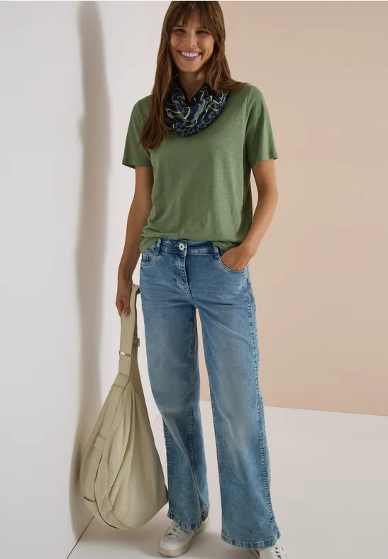 Femme en t-shirt vert, jean évasé, foulard au cou et grand sac beige sur fond neutre.