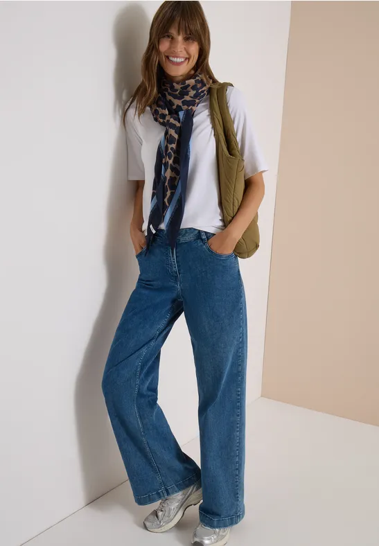 Vrouw lacht in wijdvallende jeans, wit shirt, sjaal en sneakers, poseert voor neutrale achtergrond.
