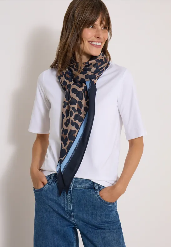 Lachende vrouw in wit shirt, patroon sjaal en jeans, handen in de zakken.