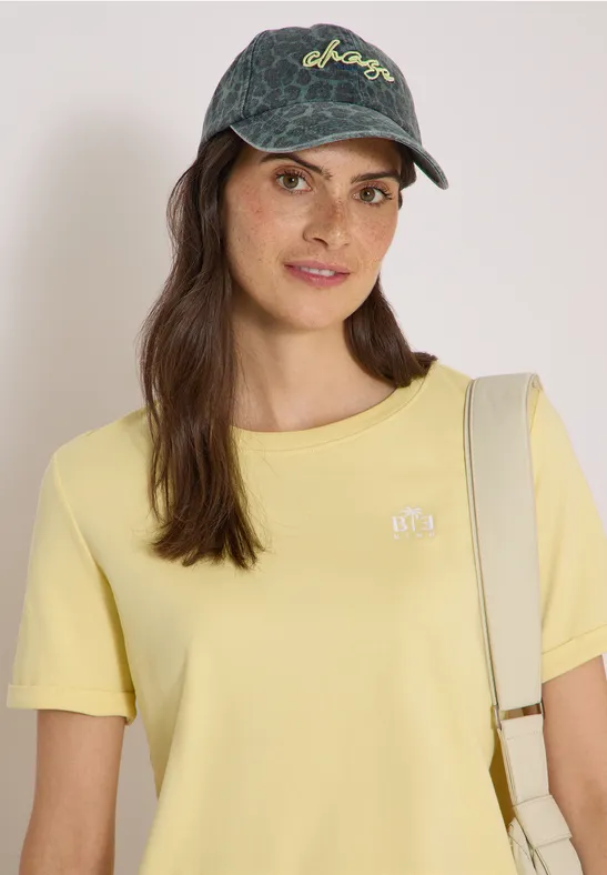 Frau mit Jeans-Cap, gelbem T-Shirt und Schultertasche, die in die Kamera lächelt.