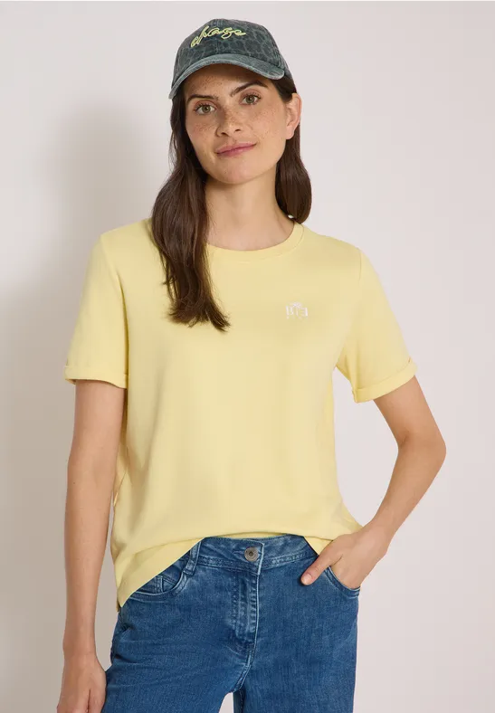 Eine Frau mit gelbem T-Shirt, blauer Jeans und Baseballcap lächelt vor neutralem Hintergrund.
