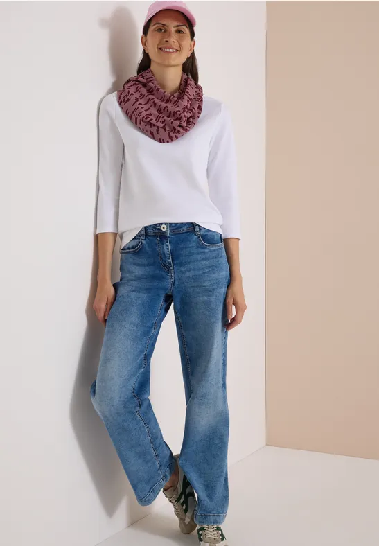 Femme en jean, haut blanc, casquette et écharpe roses, souriante, adossée à un mur clair.