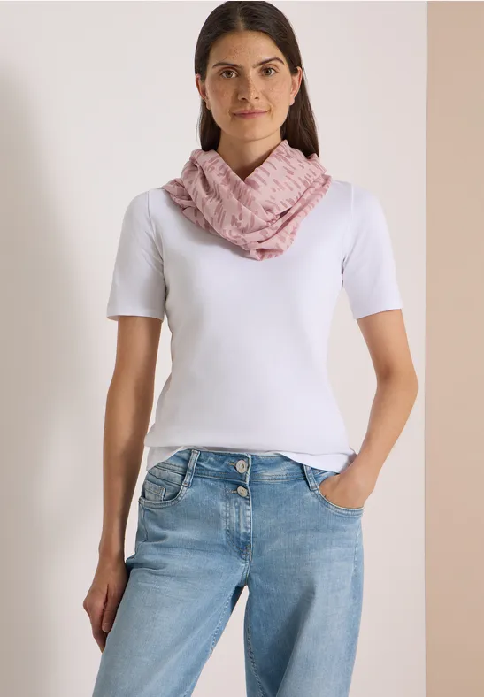 Une femme porte un t-shirt blanc, un jean et une écharpe rose, debout devant un fond clair.