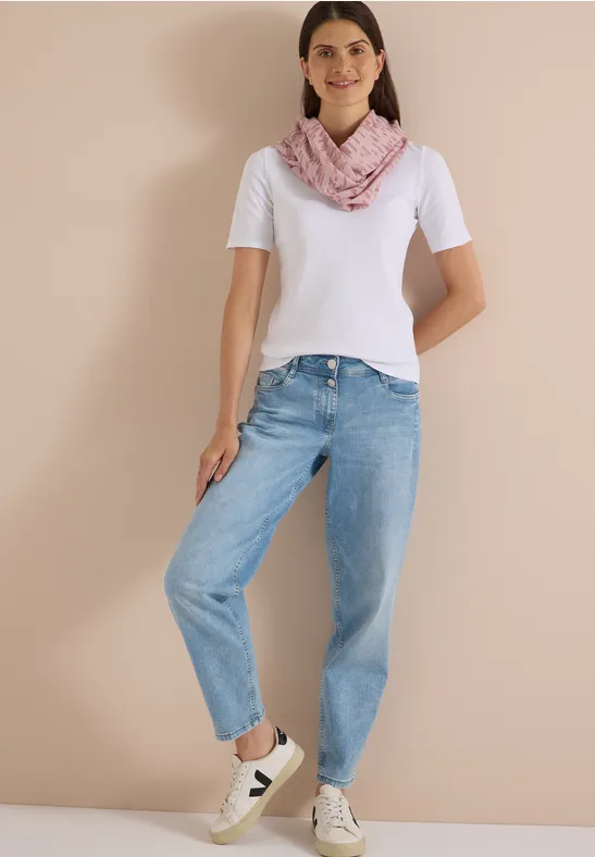 Femme en t-shirt blanc, jean bleu, foulard rose et baskets blanches, posant devant un mur neutre.