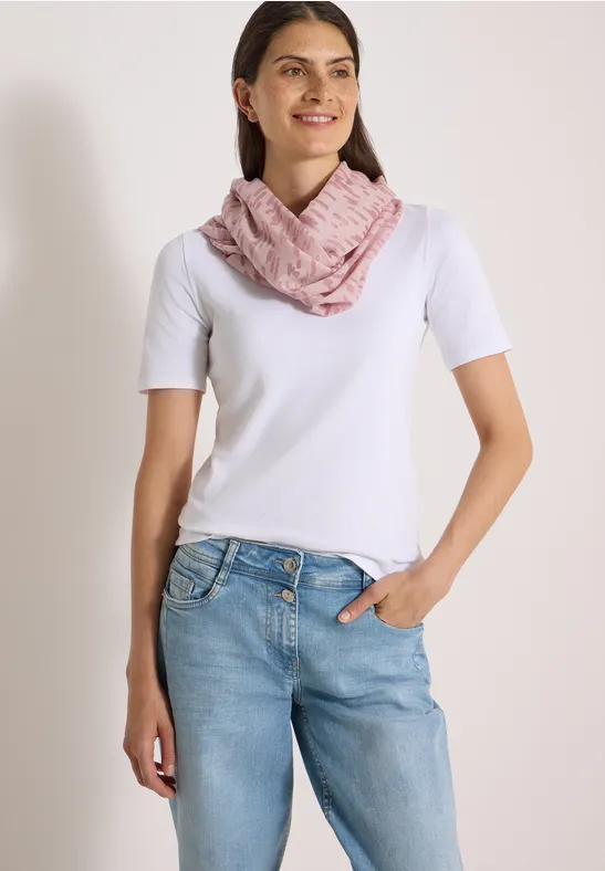 Femme en t-shirt blanc, jean bleu et écharpe rose devant un fond clair.