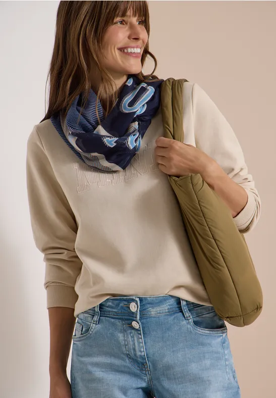 Une femme souriante en pull beige, jean, foulard bleu et sac vert rembourré.