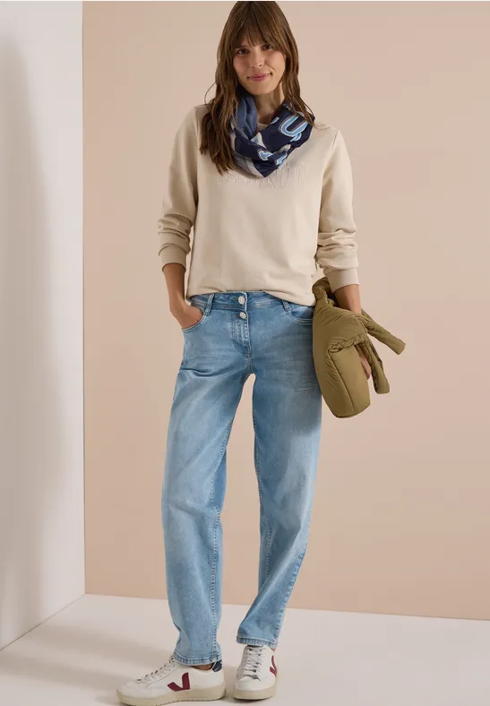 Femme en pull crème, écharpe bleue, jean ample et baskets blanches, tenant un sac vert.
