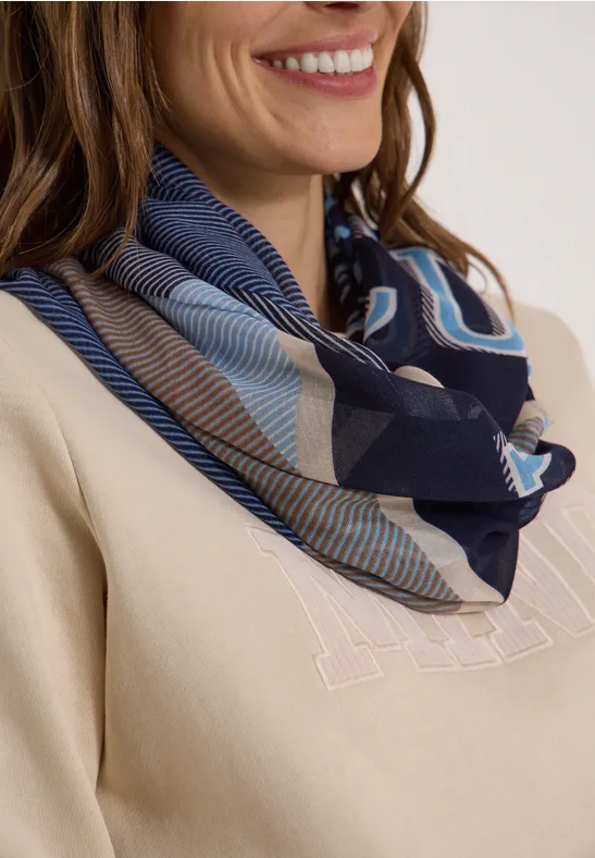 Femme avec foulard bleu à motifs autour du cou, pull beige et sourire discret.