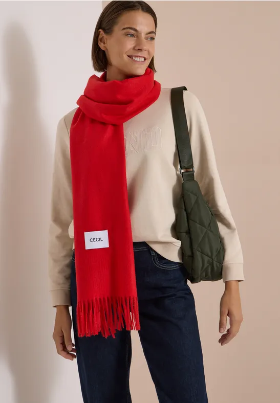 Femme souriante portant une écharpe rouge, un pull beige, un jean et un sac vert en bandoulière