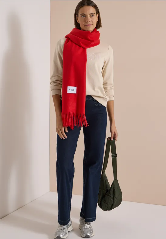 Femme en pull beige, jean bleu, écharpe rouge et sac vert.