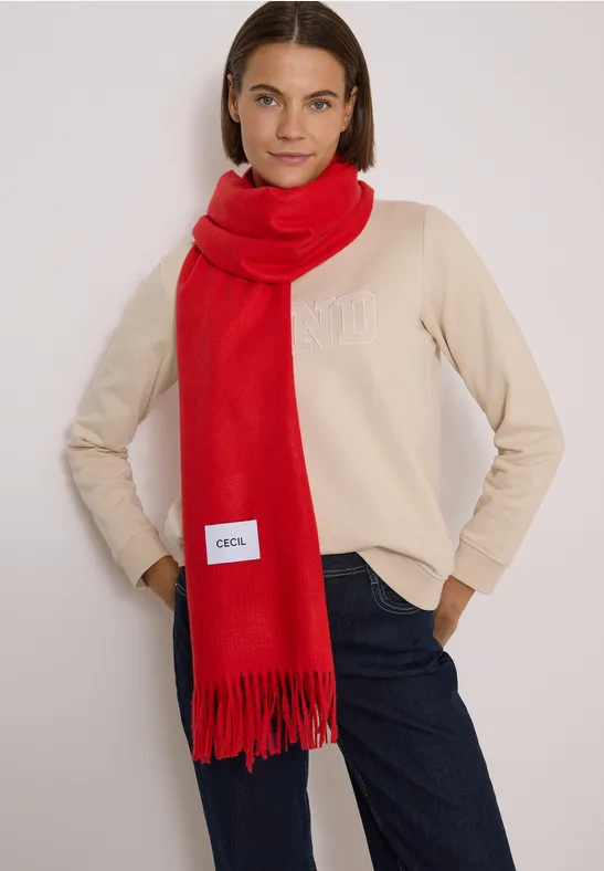 Femme portant une écharpe rouge, un pull beige et un jean sur fond neutre.