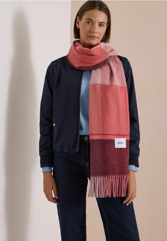 Femme en veste bleu marine, jean bleu et grand foulard rouge-rose devant un mur clair.