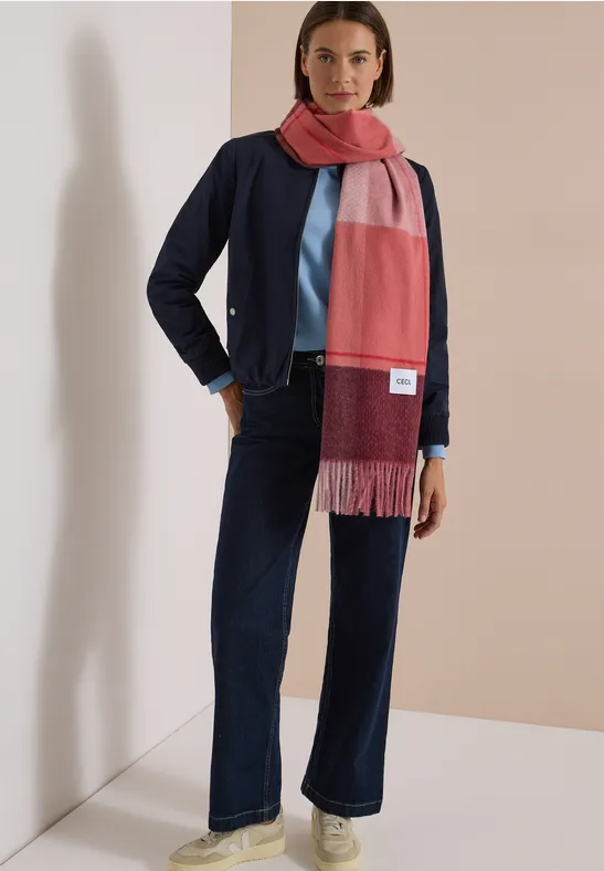 Femme portant une écharpe bicolore, veste bleu marine, haut bleu clair et jean évasé devant un fond neutre.