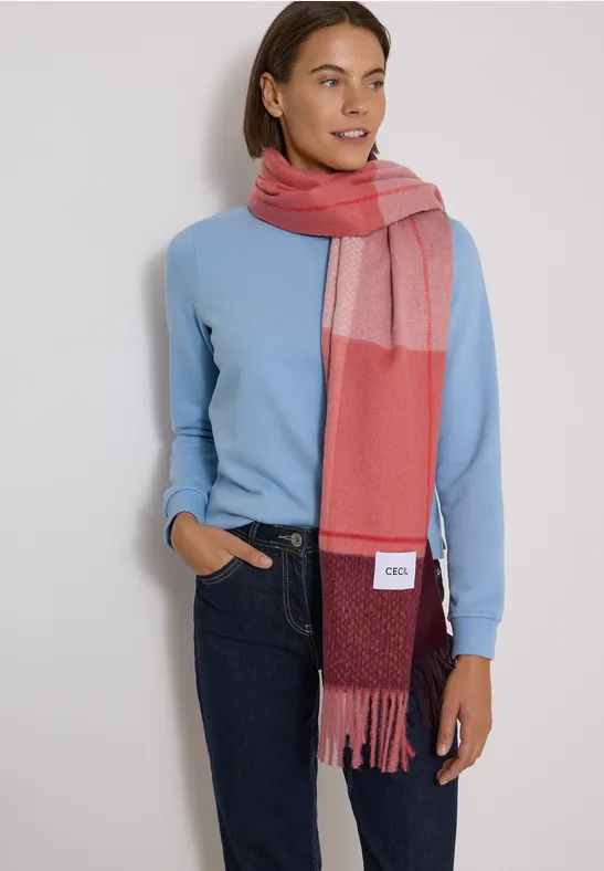 Femme souriante en pull bleu clair, jean et écharpe bicolore rouge/rose, main dans la poche.