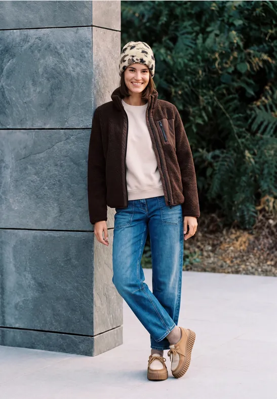 Glimlachende vrouw met muts, jas en jeans leunt bij een moderne gevel buiten.