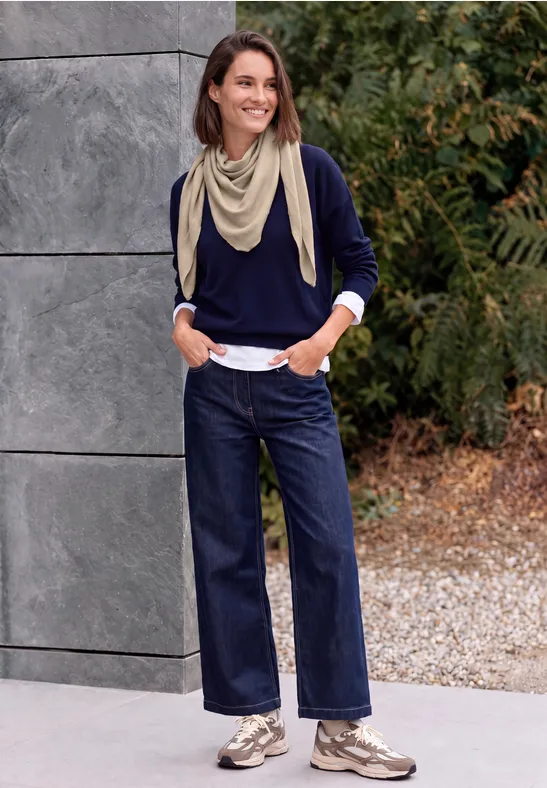 Eine Frau mit hellbeigem Schal, dunkelblauem Pullover, weiten Jeans und Sneakers vor einer Mauer mit Pflanzen.