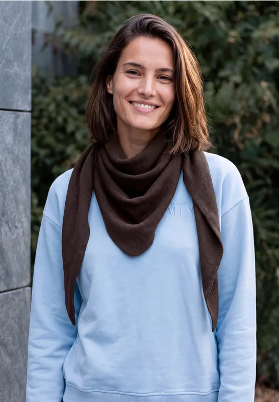 Eine lächelnde Frau in blauem Sweatshirt und braunem Schal steht vor einem grünen Hintergrund.