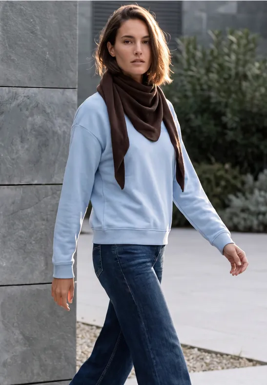 Frau in Jeans, hellblauem Pullover und dunklem Schal geht draußen an einer Betonwand vorbei.