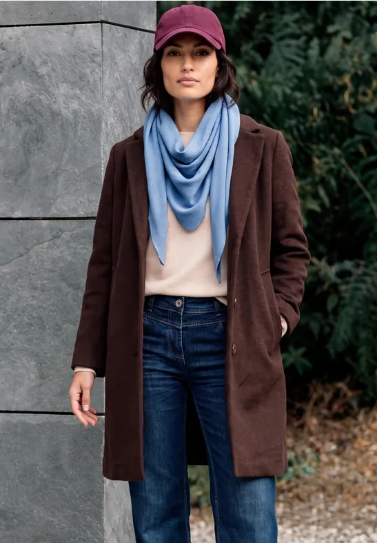 Frau mit burgunderfarbener Jacke, Jeans, rosa Pullover, blauem Schal und Cap steht vor Betonwand und Pflanzen.