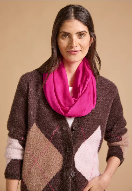 Portrait einer lächelnden Frau in braunem Strickjacke mit Rautenmuster und pinkem Schal vor beigem Hintergrund.