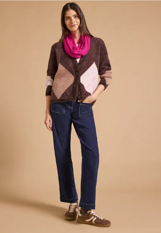 Frau trägt karierten Cardigan in Braun und Rosa, Jeans und Sneakers vor neutralem Hintergrund.