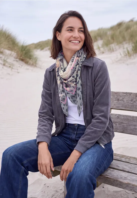 Une femme souriante assise sur un banc de plage, en jean, écharpe et veste, avec des dunes en arrière-plan.