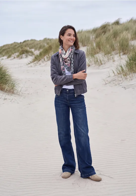 Une femme souriante en jean et foulard se tient détendue sur une plage de dunes herbeuses.